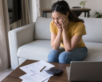 Frau sitzt mit verzweifelter Mimik am Couchtisch vor Formularen und Laptop 