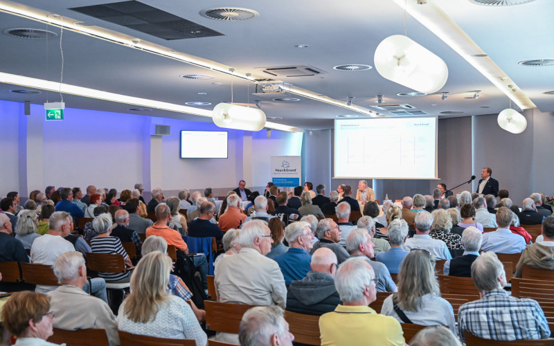 Großer Saal mit Publikum bei einer Präsentation, vorne Redner am Pult und projizierte Folien an der Wand