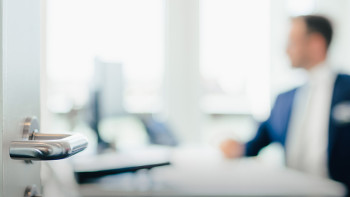 Geöffnete Glastür mit Metallgriff, im Hintergrund unscharfer Mann im Anzug an Schreibtisch in Büro.