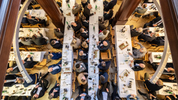 Vogelperspektive auf mehrere lange Tische mit Personen, die an einem formellen Essen in einem großen Raum mit Holzsäulen teilnehmen