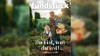 Zwei Personen stehen in einem Garten, eine trägt eine Holzkiste mit Gemüse, die andere hält einen Korb mit Kohl.