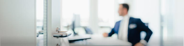 Geöffnete Glastür mit Metallgriff, im Hintergrund unscharfer Mann im Anzug an Schreibtisch in Büro.