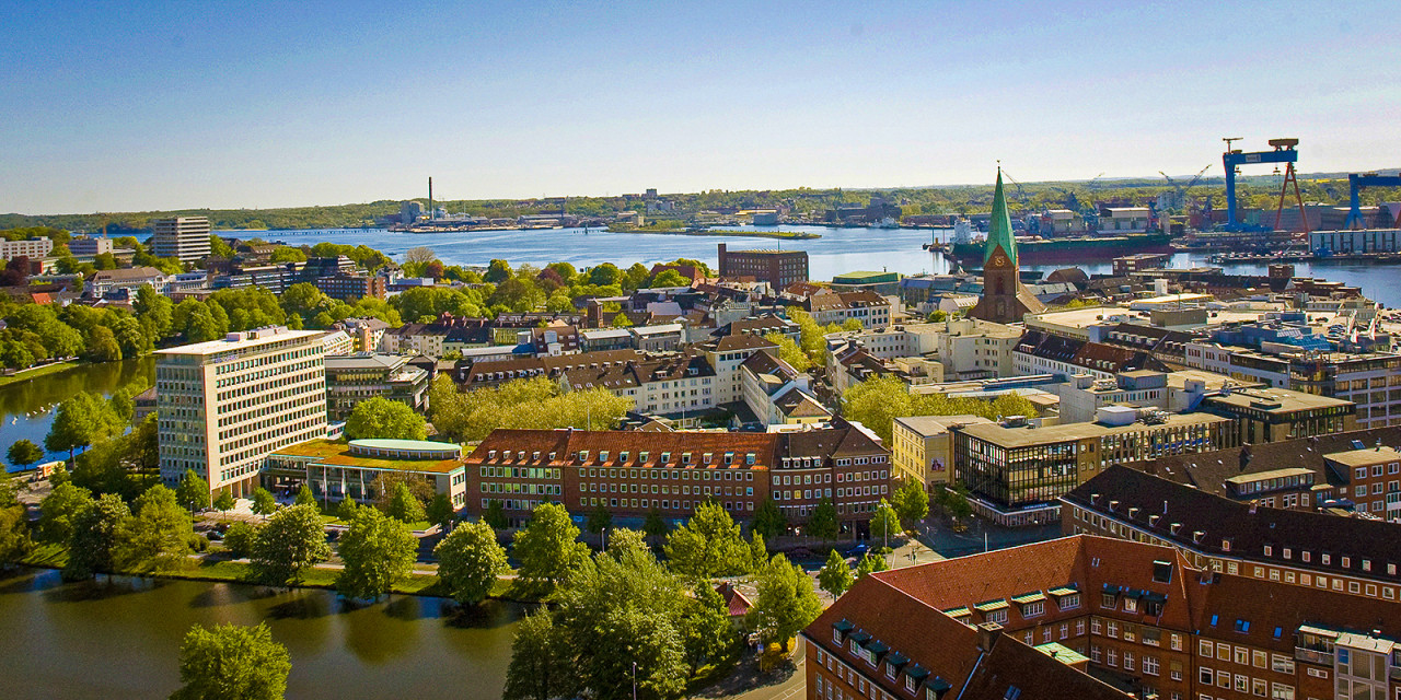 Luftaufnahme von Kiel mit Förde, mehreren Gebäuden, Bäumen und einem Hafen mit Kränen im Hintergrund.