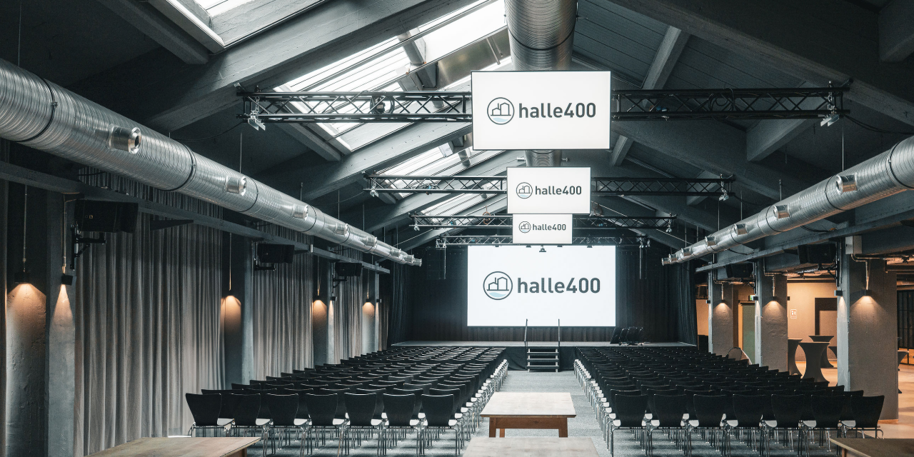 Großer Veranstaltungsraum mit Reihen von Stühlen, drei großen Bildschirmen mit dem Schriftzug 'halle400' und Tischen im Vordergrund unter einem Glasdach mit sichtbaren Lüftungsrohren