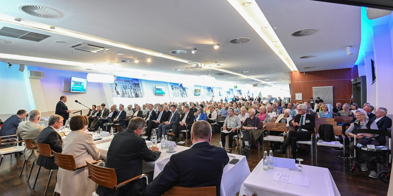 Großer Saal mit vielen sitzenden Personen bei einer formellen Veranstaltung, vorne mehrere Personen an Tischen mit Mikrofonen