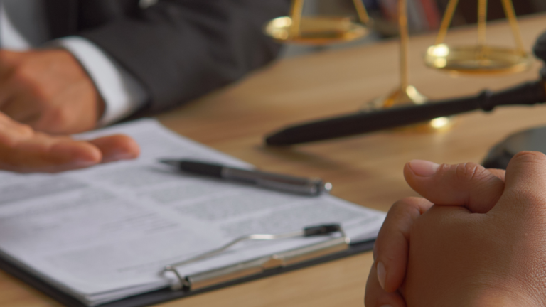 Zwei Personen sitzen an einem Tisch, eine trägt einen Anzug, auf dem Tisch liegen ein Aktenordner, ein Richterhammer und eine Waage der Justiz