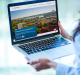 Person hält Laptop mit geöffneter Webseite, die eine Stadtansicht und Öffnungszeiten zeigt.