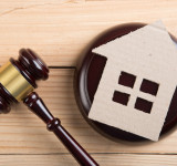 Richterhammer liegt neben rundem Holzblock mit Papphaus mit Fenstern auf hellem Holztisch.