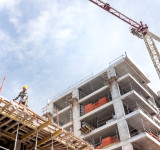 Bauarbeiter mit gelbem Helm auf einem Gerüst vor einem mehrstöckigen Rohbau und einem Baukran im Hintergrund