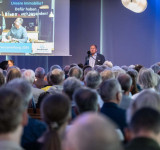 Mann spricht vor großem Publikum in einem Saal mit Präsentationsfolie im Hintergrund