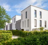 Moderne weiße Wohnhäuser mit rechteckigen Fenstern umgeben von grünen Büschen und Bäumen unter blauem Himmel