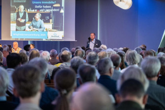 Mann spricht vor großem Publikum in einem Saal mit Präsentationsfolie im Hintergrund