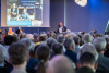 Mann spricht vor großem Publikum in einem Saal mit Präsentationsfolie im Hintergrund