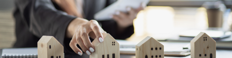 Person platziert kleine Holzhäuser auf einem Tisch und symbolisiert damit Immobilienverwaltung, Hauskauf oder Wohnungsplanung im Büroalltag.